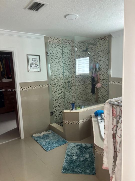 Walk-in glass-enclosed shower featuring a mosaic-tiled accent wall, built-in soaking tub with decorative tile border, and large-format neutral-tone tile flooring
