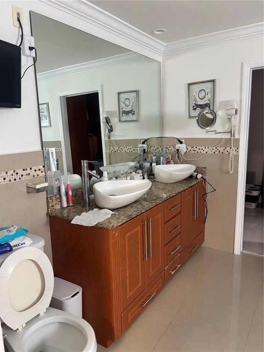 Dual vanity featuring oval vessel sinks, stone-finish countertops, warm-toned wood cabinetry with metallic pulls, and a mosaic tile backsplash