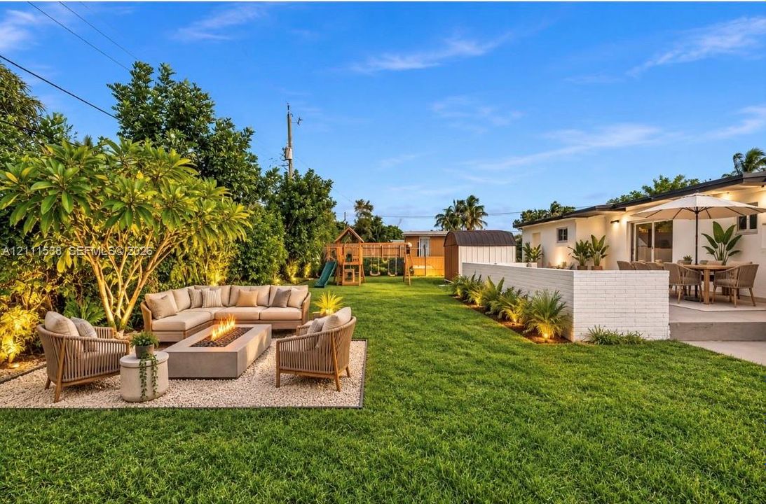 Expansive backyard featuring a manicured lawn, stone-surfaced patio area, painted brick perimeter wall, and mature landscaping