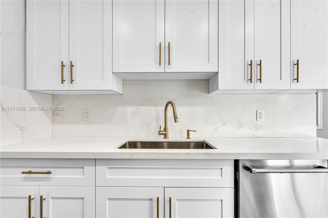 Shaker-style white cabinetry with metallic pulls, white stone-finish countertops, full-height stone-finish backsplash, undermount stainless steel sink with a high-arc metallic faucet, and stainless steel dishwasher
