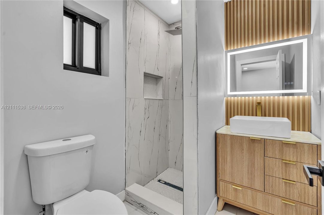 Bathroom featuring a wood-finish vanity with a rectangular vessel sink, LED-backlit mirror, vertical wood-slat accent wall, and a walk-in shower with large-format tile surrounds and a built-in niche