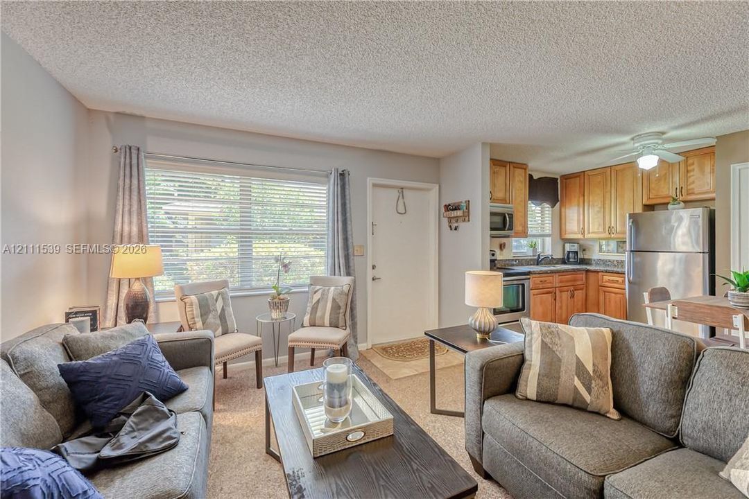 Open-concept interior featuring wood-finish cabinetry, stainless steel appliances, neutral wall tones, carpeted flooring, and a large horizontal window