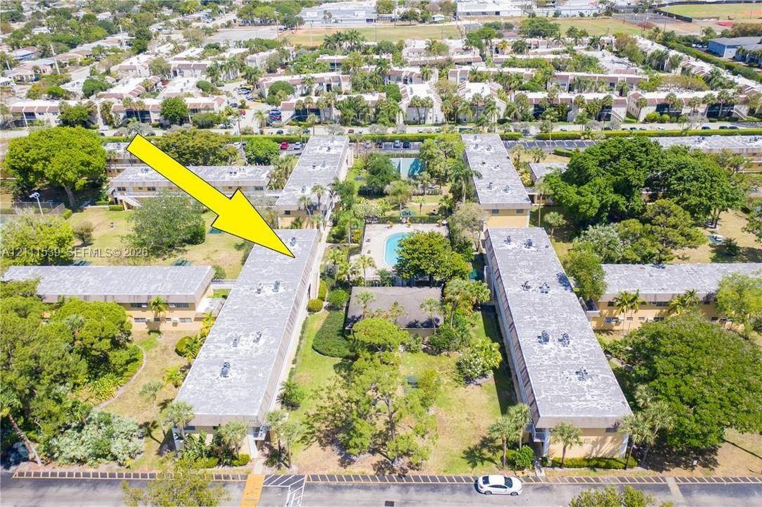 Aerial perspective of the residential complex featuring multi-story structures, integrated swimming pool, expansive landscaped grounds, and mature palm trees