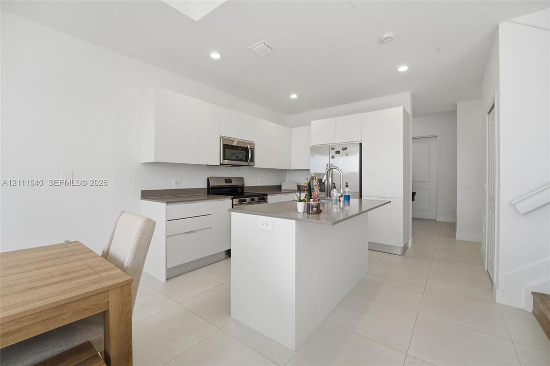 Kitchen featuring flat-panel white cabinetry, stone-surface countertops, a center island with integrated sink, stainless steel appliances, and large-format tile flooring