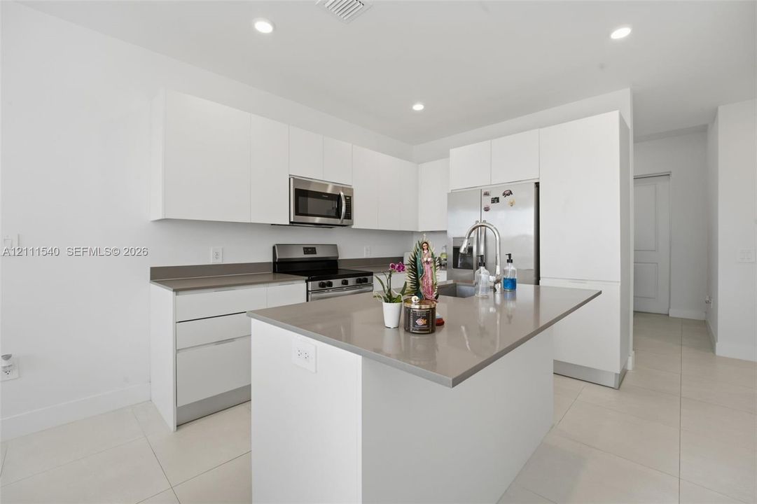 Kitchen featuring flat-panel white cabinetry, solid-surface countertops, a center island with integrated sink, stainless steel appliances, and large-format tile flooring