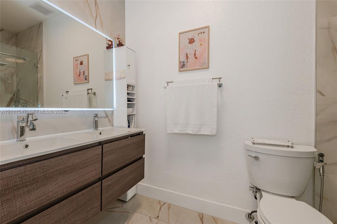 Double-vanity with a wood-finish floating cabinet, large LED-backlit mirror, integrated sink basins, polished metal faucets, and large-format tile flooring