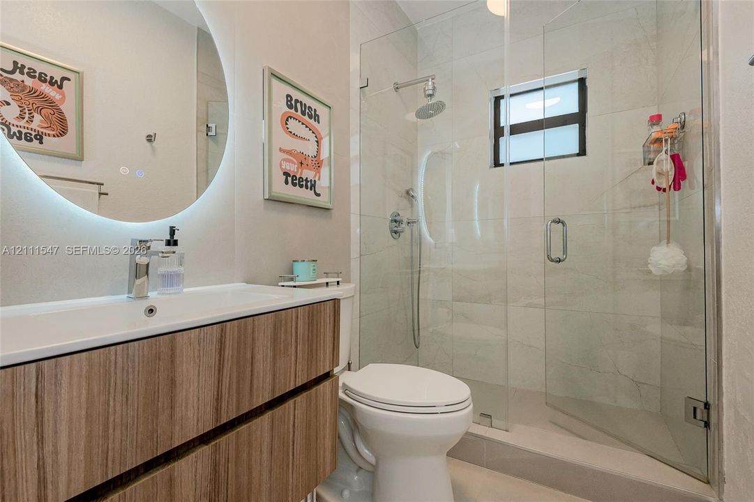 Floating wood-finish vanity with integrated sink, circular backlit mirror, and glass-enclosed shower featuring tiled surround and rainfall showerhead