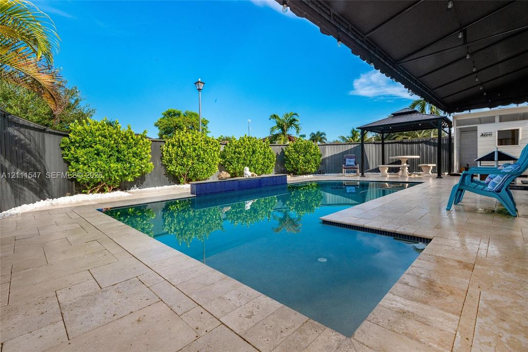 Swimming pool featuring a raised tiled spillway, expansive stone-paver decking, black metal-frame gazebo, and perimeter privacy fencing