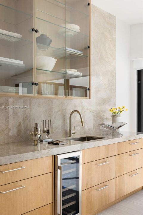 Wet bar featuring light-toned wood cabinetry, stone slab backsplash and countertop, under-counter wine refrigerator, and upper glass-front cabinetry with interior shelving