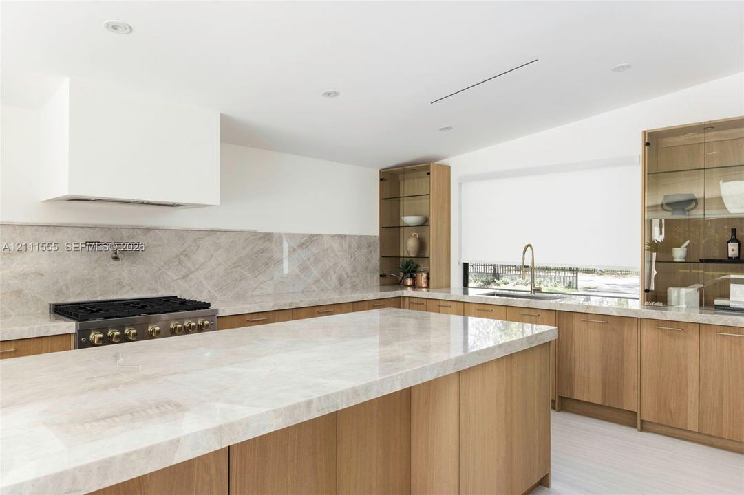 Kitchen featuring natural wood cabinetry, stone-surface countertops and backsplash, integrated glass-front display shelving, wall-mounted pot filler, and a large central island