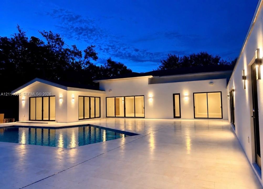 Contemporary exterior featuring smooth stucco finish, expansive stone patio, integrated wall-mounted lighting, multiple sliding glass door sets, and a rectangular swimming pool
