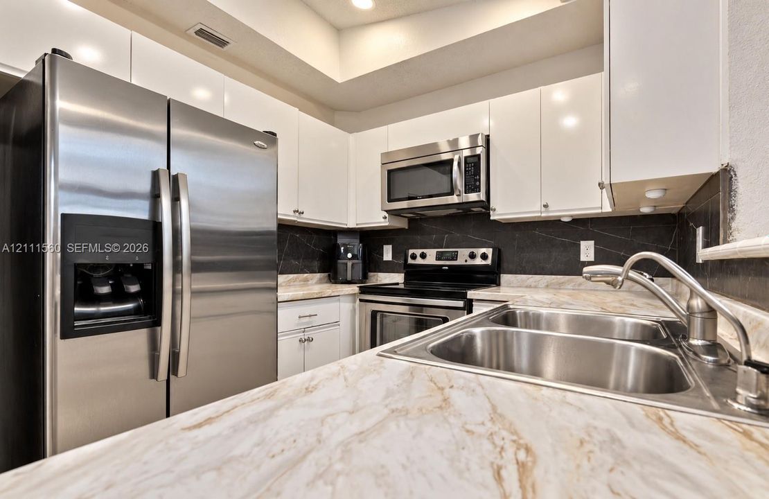Kitchen featuring white gloss cabinetry, stone-patterned countertops, stainless steel appliances, and a dark stone-finish backsplash with an integrated dual-basin stainless steel sink
