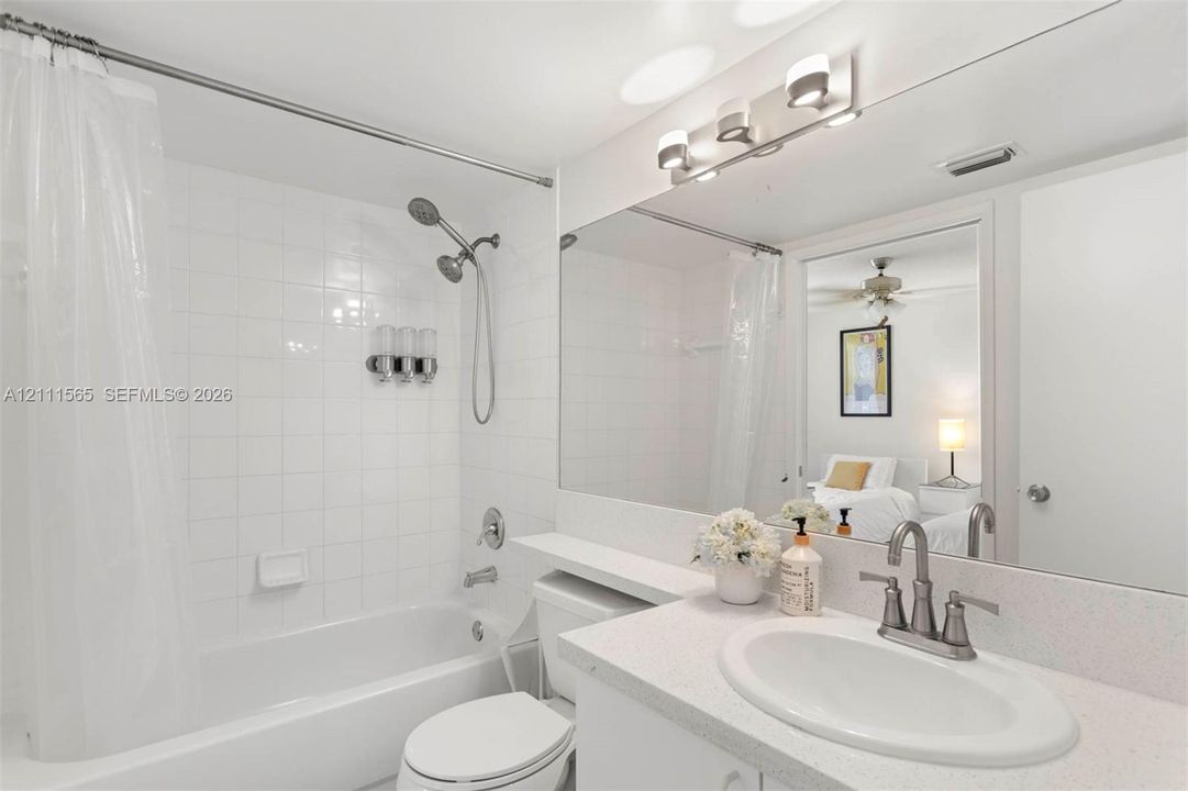 Bathroom featuring a white vanity with a composite countertop, an integrated oval sink, a brushed-metal widespread faucet, a large frameless mirror, and a shower-tub combination with white tile surround