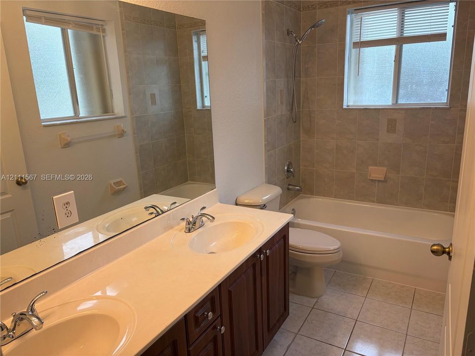 Double-sink vanity with a solid-surface countertop, dark wood-finish cabinetry, ceramic tile flooring, and a shower-tub combination featuring full-height wall tiling