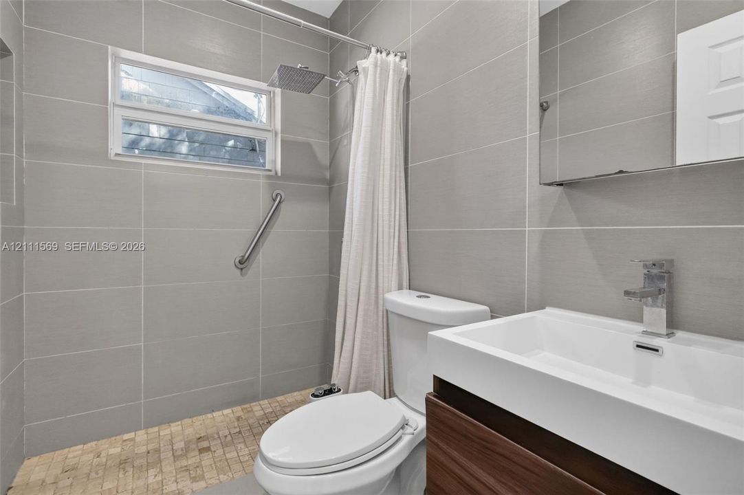 Bathroom featuring large-format gray wall tile, wood-finish vanity with an integrated rectangular sink, wall-mounted rain shower head, and textured stone floor tile