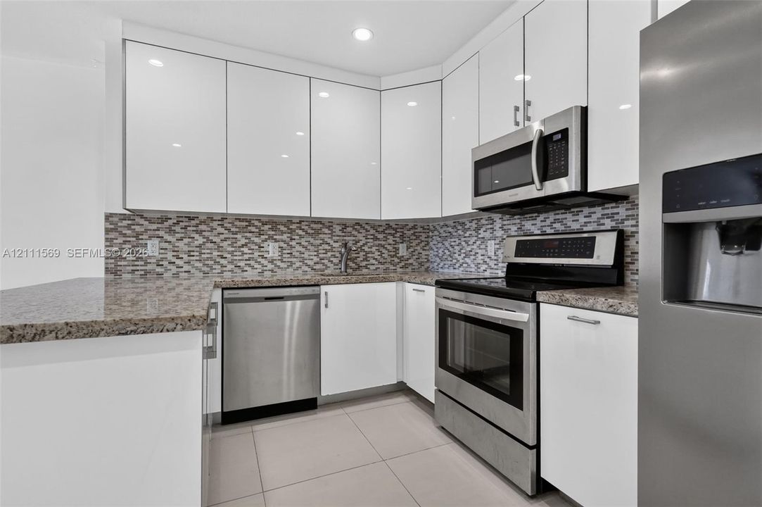Kitchen featuring high-gloss white cabinetry, stone-finish countertops, stainless steel appliances, and a glass-tile mosaic backsplash