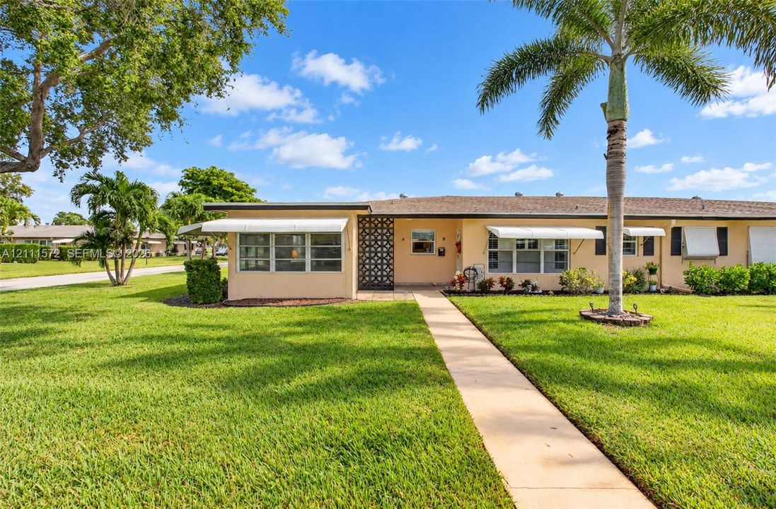 Single-story corner unit villa, metal window awnings, concrete breezeway blocked patio, and established landscaping with palm trees