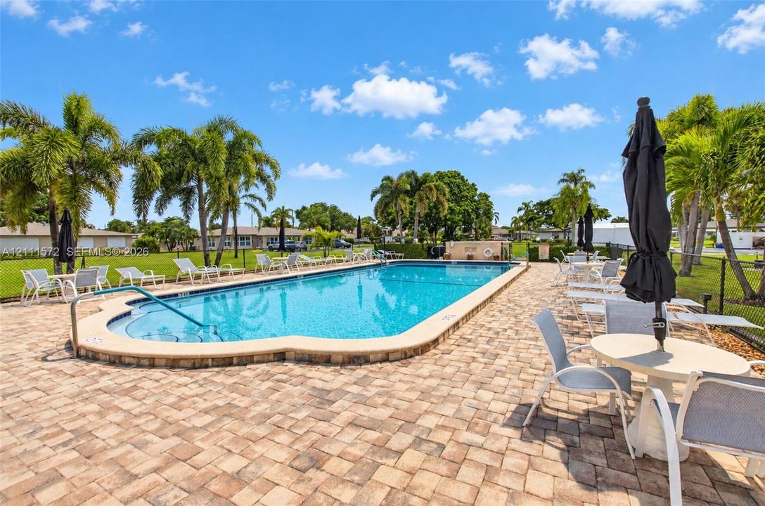 In-ground pool featuring a curved entry step, metal handrail, paver stone decking, and surrounding palm trees