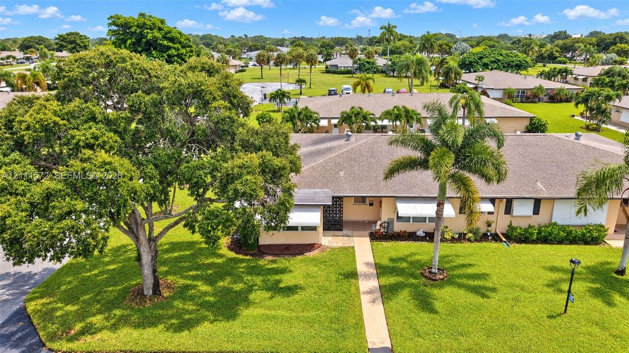 Single-story corner residence featuring a shingled roof, exterior shades, concrete walkway, mature landscaping, and an expansive lawn in a well maintained, friendly community