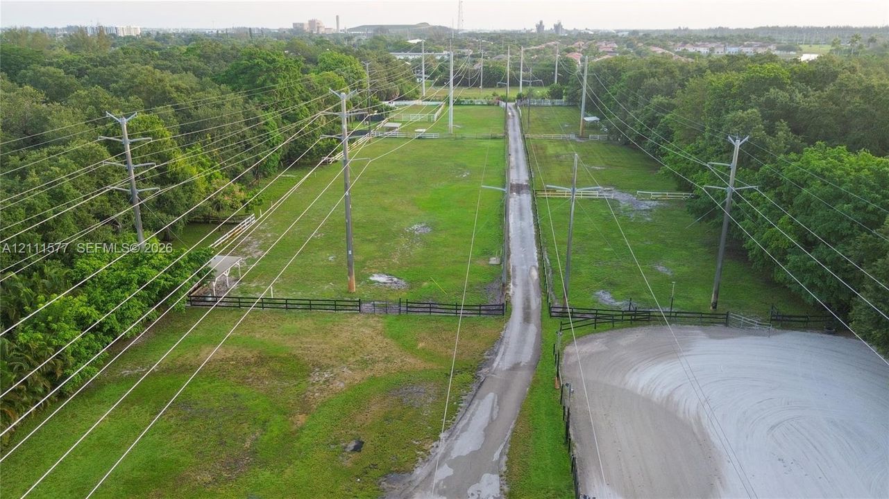 Expansive grounds featuring a central paved driveway, perimeter post-and-rail fencing, a dedicated sand ring, and mature tree buffers