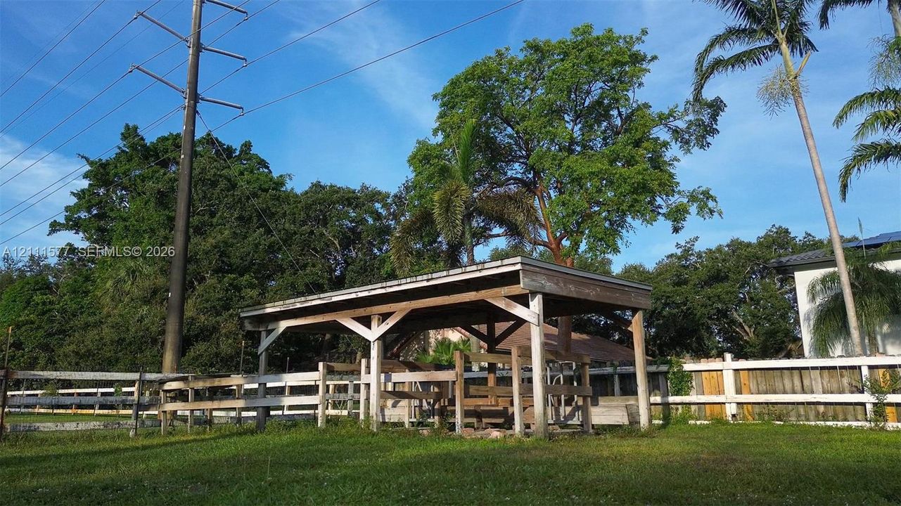 Open-air wood structure featuring a post-and-beam frame, set within a fenced area with mature trees