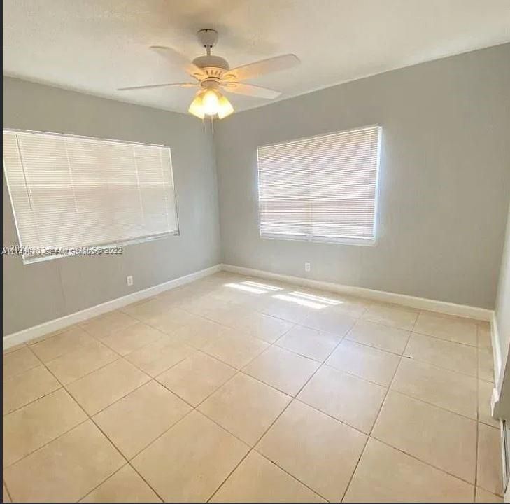 Neutral-toned walls featuring large-format tile flooring, a multi-blade ceiling fan with integrated light fixture, and two windows with horizontal blinds