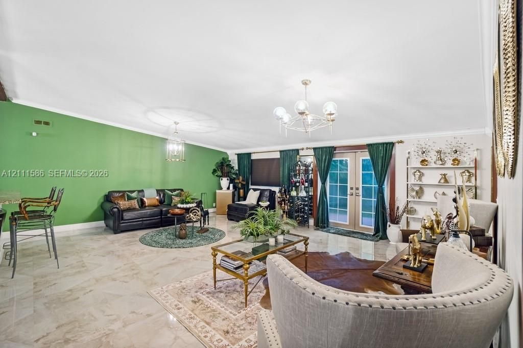 Open-concept living space featuring high vaulted ceilings, light-toned stone-finish flooring, a multi-arm polished metal chandelier, and full-height glass-paneled double doors