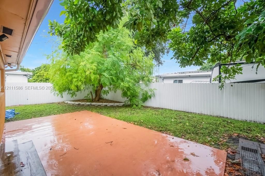 Rear exterior space featuring a concrete patio, perimeter vinyl fencing, and a mature tree within a landscaped garden bed