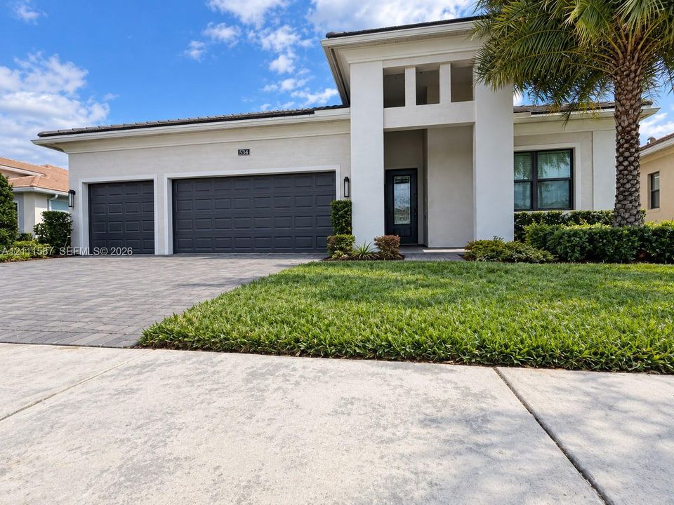 Contemporary exterior featuring a stucco facade, three-car garage with dark-toned paneled doors, paver driveway, and a recessed entryway with vertical architectural columns