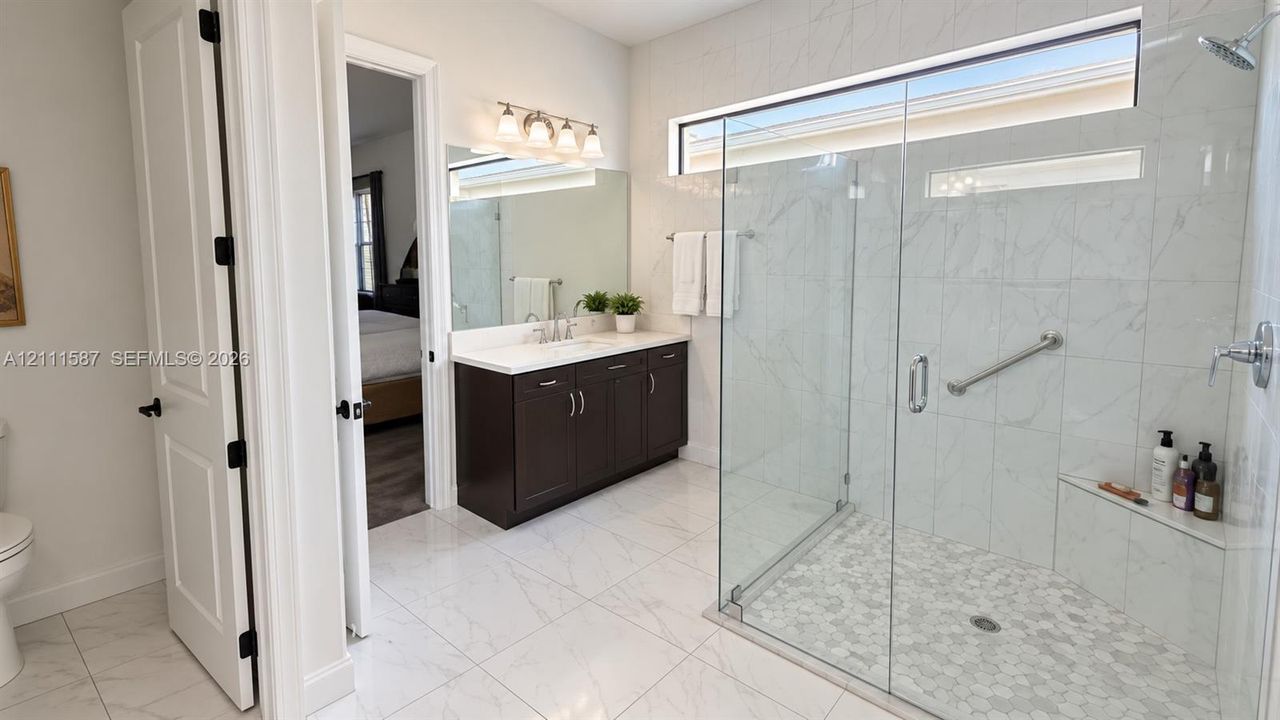 Walk-in shower with stone-finish tile surround, frameless glass enclosure, and built-in bench seating, complemented by a dark wood vanity with white countertop and large vanity mirror