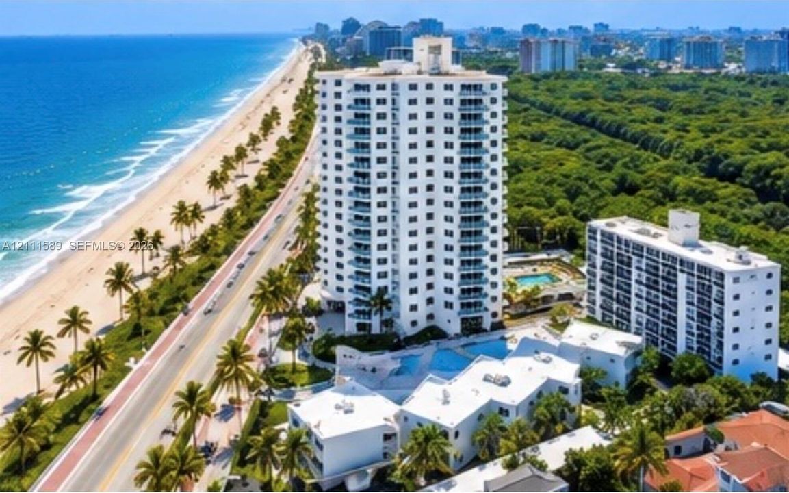 High-rise residential building featuring exterior balconies, a ground-level swimming pool, lush landscaping, and coastal frontage