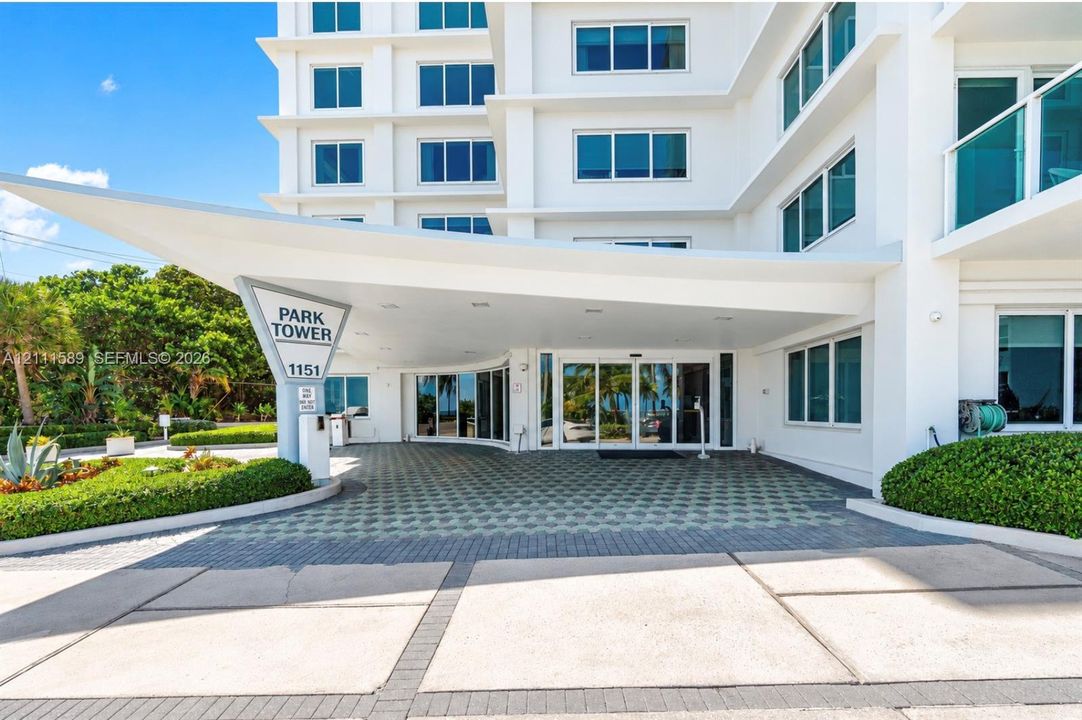 Modern high-rise exterior featuring a cantilevered covered entryway, glass sliding doors, concrete and paver drive, and manicured perimeter landscaping