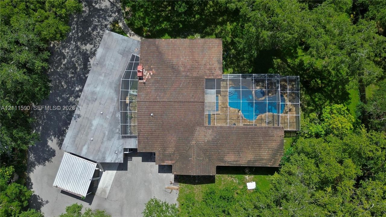 Aerial perspective featuring a multi-level roofline, screened-in swimming pool enclosure, detached covered parking structure, and mature trees on a wooded lot