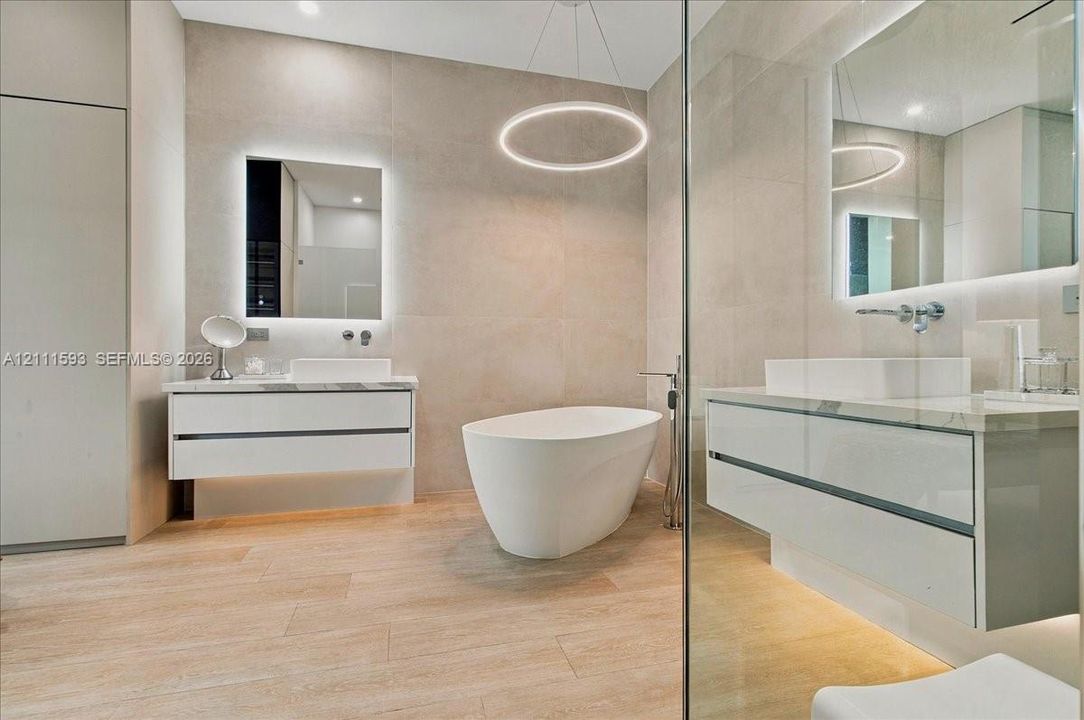Freestanding soaking tub, dual floating vanities with rectangular vessel sinks, floor-to-ceiling tile walls, wood-finish flooring, and minimalist circular pendant lighting