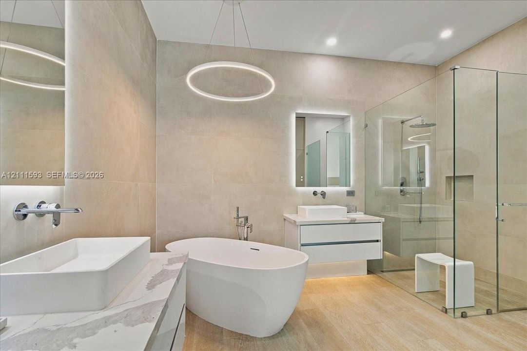 Freestanding soaking tub, floating vanity with vessel sink, walk-in glass-enclosed shower, wall-mounted faucets, and recessed circular LED pendant lighting