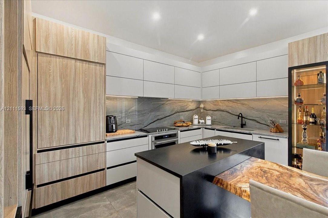Contemporary kitchen featuring matte finish cabinetry, expansive natural stone backsplash, integrated stainless steel appliances, a center island with a stone surface and wood-finish accent extension, and a built-in illuminated glass display cabinet