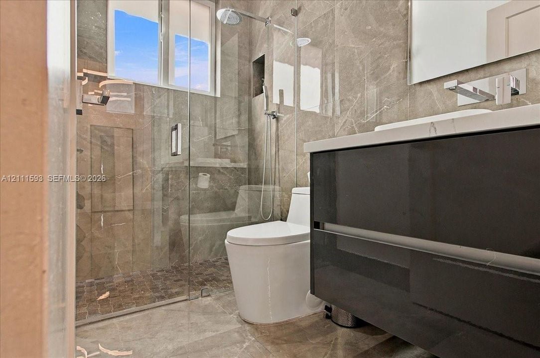Bathroom featuring full-height stone tile walls, glass-enclosed shower with rainfall showerhead, floating charcoal-finish vanity, and wall-mounted chrome faucet