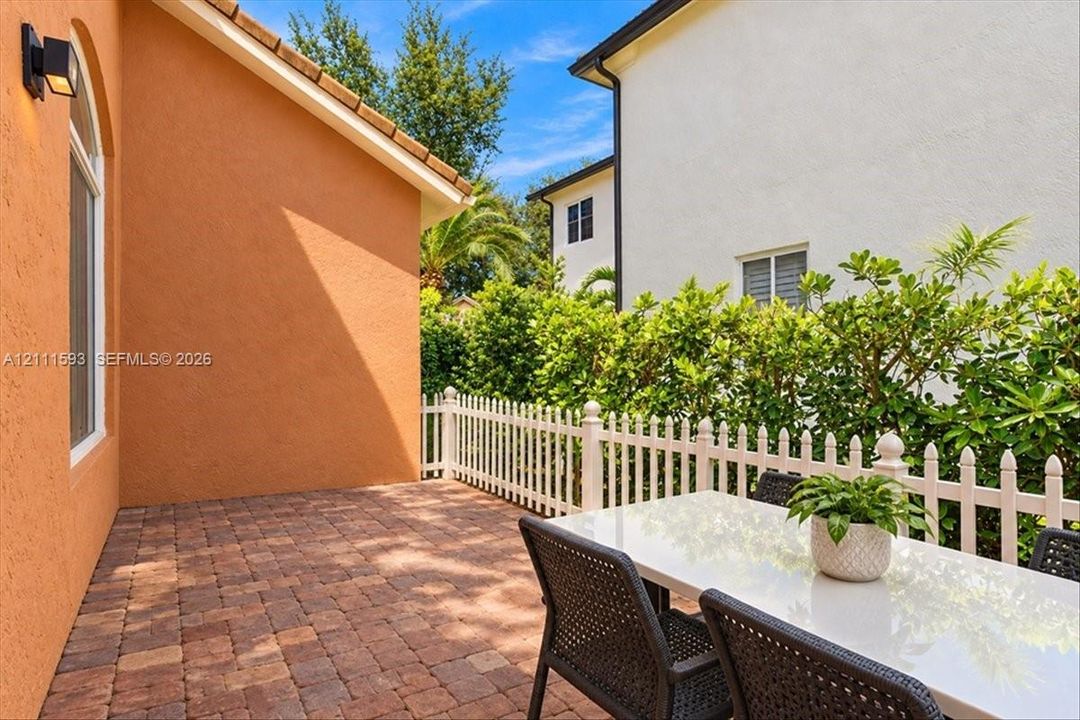 Paved side patio featuring stone-finish pavers, white picket fencing, and exterior wall-mounted lighting