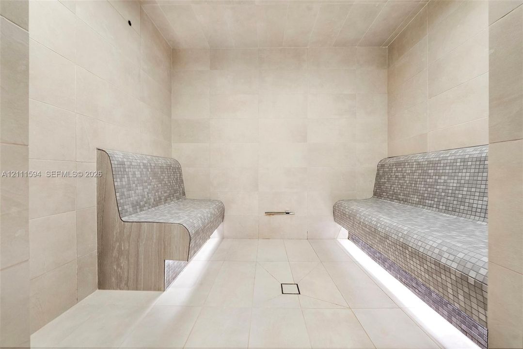 Steam room featuring large-format stone wall and floor tile, built-in dual benches with mosaic tile surfacing, integrated under-bench lighting, and a centered floor drain