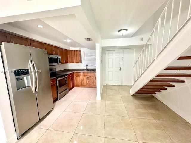 Kitchen featuring wood cabinetry, stainless steel appliances, and stone-finish countertops, paired with a staircase with wood-finish treads and tile flooring