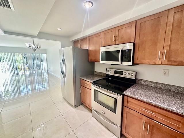 Kitchen featuring natural wood cabinetry, speckled stone countertops, stainless steel appliance suite, and light-toned tile flooring