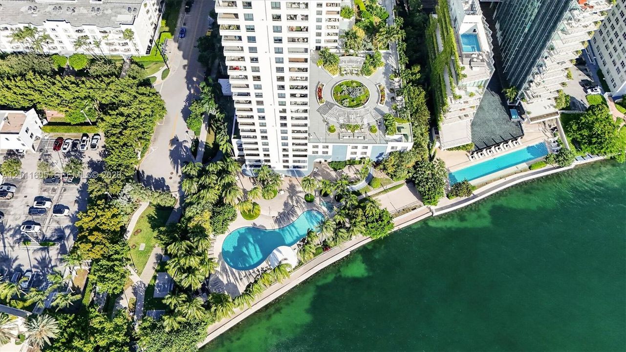 Aerial perspective of a high-rise residential building featuring a landscaped podium terrace with a circular water feature, a kidney-shaped ground-level swimming pool, and direct proximity to the waterfront
