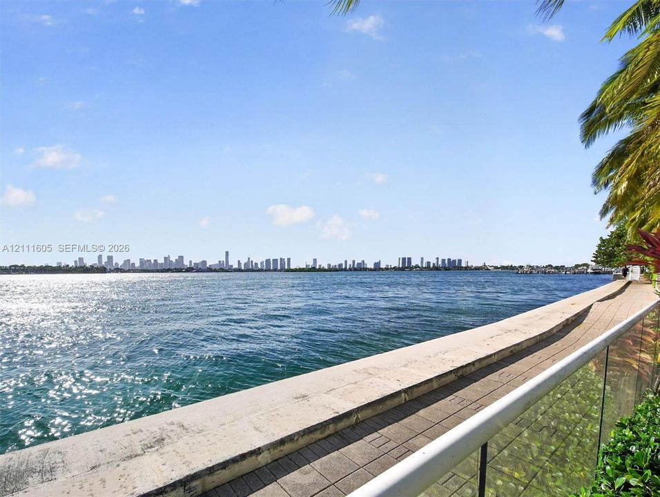 Waterfront property featuring a concrete seawall, paver-stone walkway, glass-panel railing, and tropical palm foliage