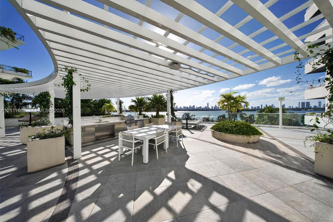 Expansive rooftop terrace featuring a curved white pergola, built-in stainless steel outdoor kitchen, stone tile flooring, and integrated concrete planters