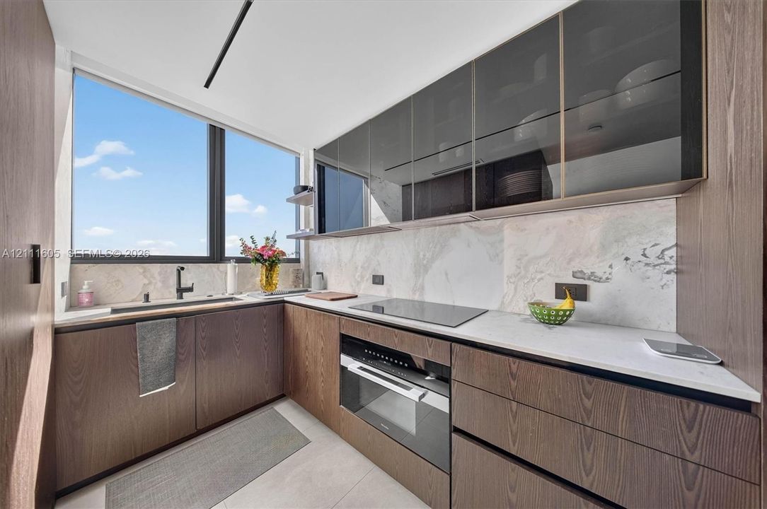 Kitchen featuring wood-finish cabinetry, stone-patterned backsplash, light-colored countertops, integrated cooktop and oven, and upper glass-front cabinets