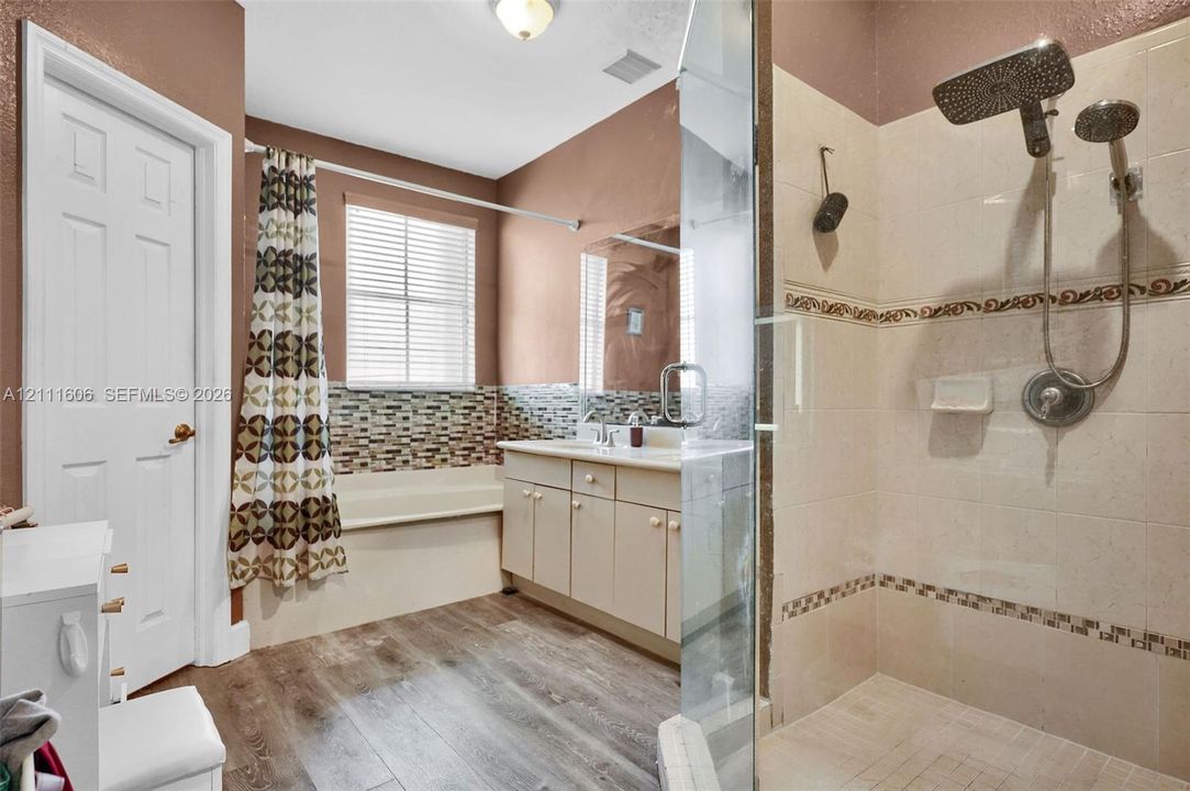 Bathroom featuring wood-finish flooring, a glass-enclosed shower with a rainfall showerhead, a built-in tub with decorative tile backsplash, and a vanity with a mirror