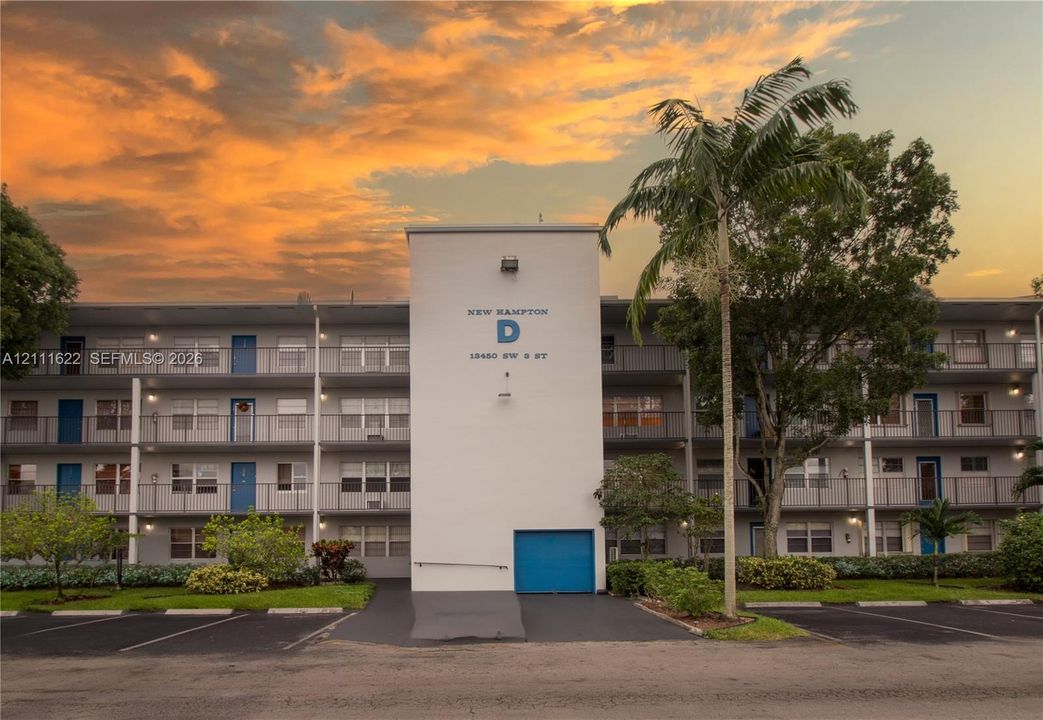 Three-story multi-unit structure featuring a smooth-finish exterior, covered balcony railings on every level, paved surface parking, and mature palm landscaping