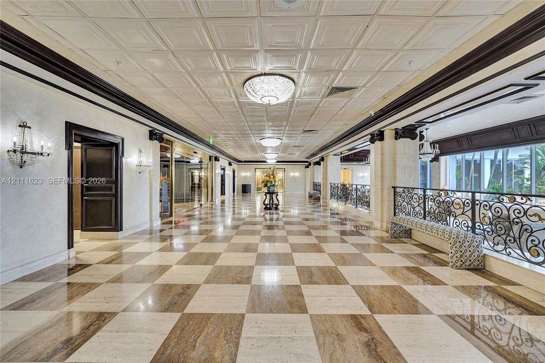 Grand gallery featuring a coffered ceiling, polished stone checkerboard flooring, ornate iron railings, custom crown molding, and crystal-style chandelier lighting