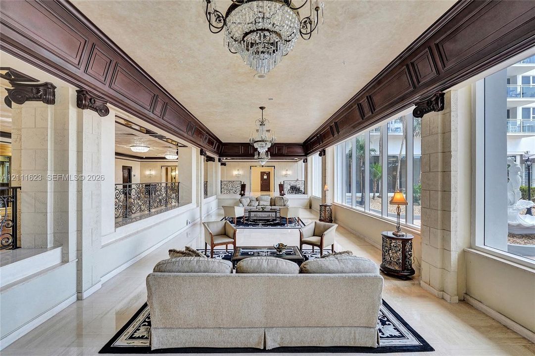 Expansive hall featuring ornate dark wood crown molding, stone-textured pillars, floor-to-ceiling windows, polished stone flooring, and tiered crystal chandeliers