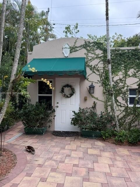 Stucco exterior featuring a scalloped green fabric awning, paneled entry door, wall-mounted exterior lantern, and patterned paver driveway